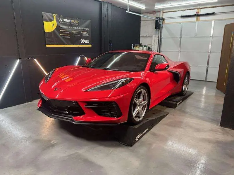 Red C8 Corvette Stingray on ramps in the Great Lakes Clear Bra install bay under an XPEL Ultimate Plus banner
