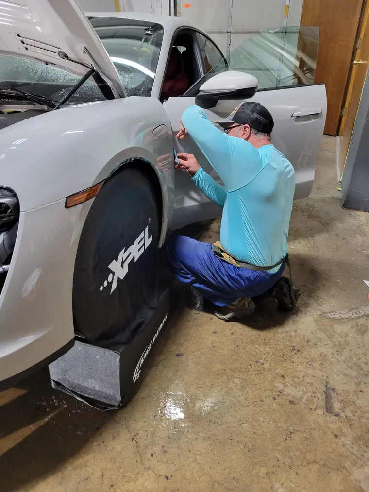 Installer fitting paint protection film along the rear quarter panel of a white Porsche, with an XPEL fender cover on the wheel